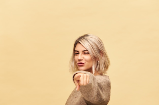 Free photo cute charming young blonde woman in cashmere sweater reaching out hand and pointing index finer, choosing you, inviting to dance with her, having energetic enthusiastic look, smiling