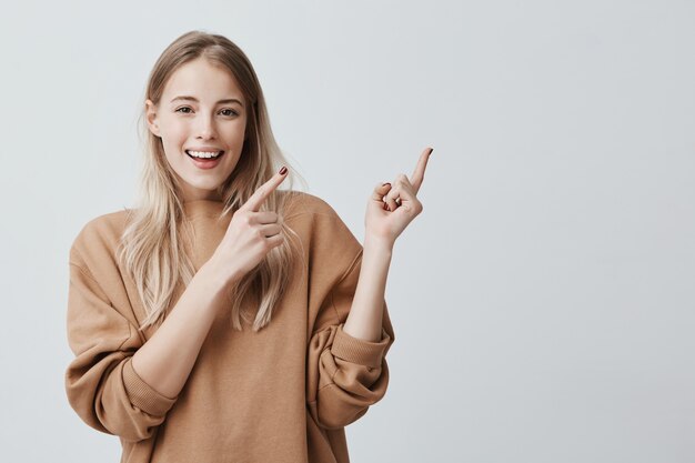 귀여운 쾌활한 금발의 젊은 여성 광범위하게 웃고 멀리 손가락을 가리키는, 재미 있고 흥미로운 것을 보여주는