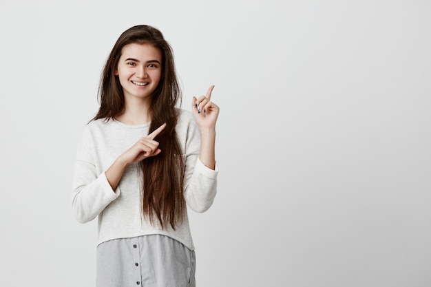 Cute cheerful brunette young woman pointing fingers away, showing something interesting and exciting