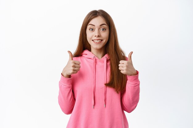 Cute cheerful girl shows thumbs up to like and approve your choice, nod and say yes, give positive feedback, recommends product, white wall