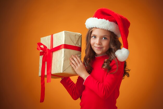 The cute cheerful little girl with Santa hat and gift on orange background