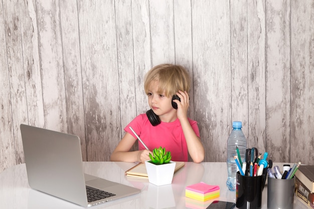 cute child boy in pink t-shirt black earphones using grey laptop on the table along with green plant bottle pens on the grey wall
