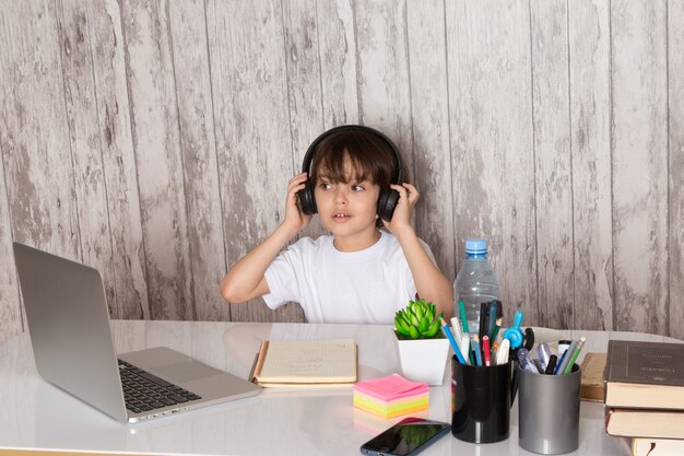 cute child boy in white t-shirt black earphones using grey laptop on the table along with green plant