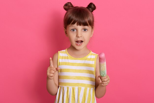 Free photo cute child girl holds ice cream keeps mouth opened, charming girl eats tasteless dessert while posing isolated on pink