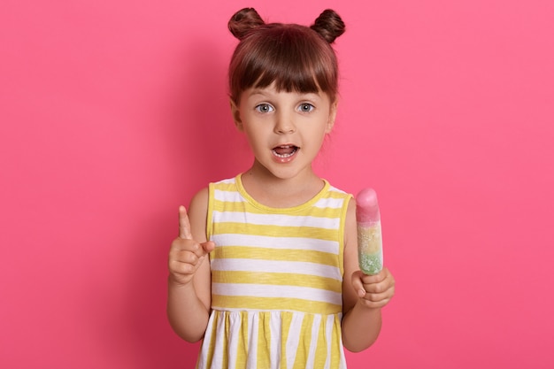 귀여운 아이 소녀 보유 아이스크림 입을 열어 유지, 매력적인 소녀 핑크에 고립 된 포즈를 취하는 동안 맛없는 디저트를 먹는다