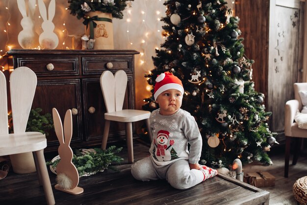 クリスマスツリーの近くの木の床に座っているサンタの帽子とパジャマのかわいい子供