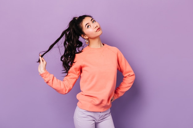 Cute chinese young woman playing with hair. Studio shot of adorable asian woman standing with hand on hip isolated on purple background.