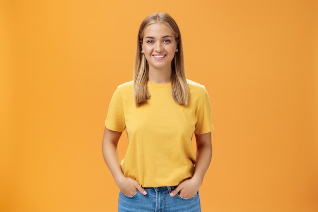 Cute chubby female with tanned skin and fair hair posing optimistic and joyful against orange background holding hands in pockets smiling broadly at camera charismatic and friendly-looking