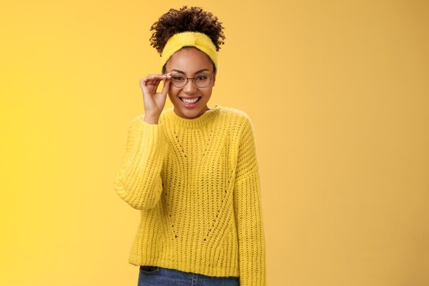 귀엽고 영리한 대학생 여대생 아프리카계 미국인 터치 안경 코가 활짝 웃으며 아르바이트 교사 어린이 영어 수업을 하며 친근한 노란색 배경 스웨터, 머리띠를 하고 있습니다.