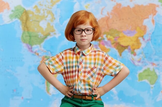 Cute confident little boy with ginger bob hairstyle wearing eyeglasses holding hands on his waist, posing against world map. Childhood, learning and education