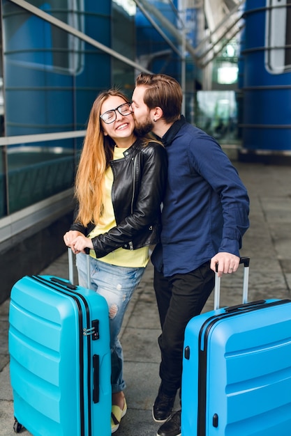 Cute couple with suitcases is standing outside in airport. She has long hair, glasses,  yellow sweater, jacket. He wears black shirt, beard. Guy is hugging and kissing the girl.