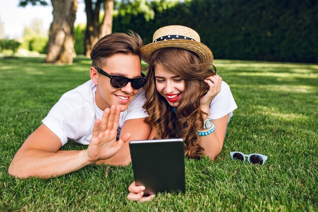 Cute couple of young people is lying on grass in summer park. Girl in hat with long curly hair holds tablet, they have good mood and communicate on tablet.