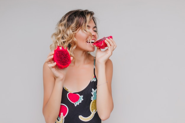 Cute curly girl eating pitaya with pleasure. Indoor photo of fair-haired caucasian woman enjoying exotic fruit.