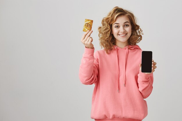 Cute curly-haired girl showing golden credit card and mobile phone screen