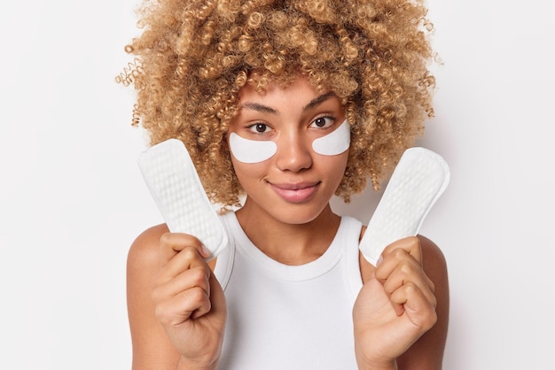 かわいい縮れ毛の女性は、月経用の生理用ナプキンを保持し、皮膚治療のために目の下にパッドを着用し、白い背景の上に分離されたカジュアルなTシャツを着用します。重要な日の衛生的な製品