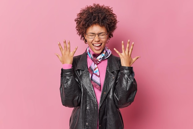 Free photo cute curly haired young woman raises hands exclaims loudly wears leather jacket spectacles kerchief around neck feels very emotionally isolated over pink background shakes palms shows emotions