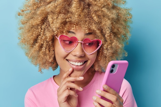 Foto gratuita carina giovane donna dai capelli ricci indossa eleganti occhiali da sole rosa a forma di cuore e maglietta tiene chat sul cellulare con il fidanzato guarda video isolato su sfondo blu tecnologie moderne