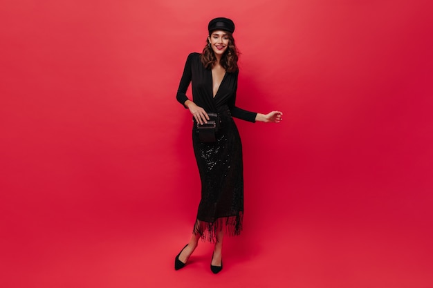 Cute curly woman in shiny skirt, black blouse and cap holds clutch bag, smiles and poses on red wall