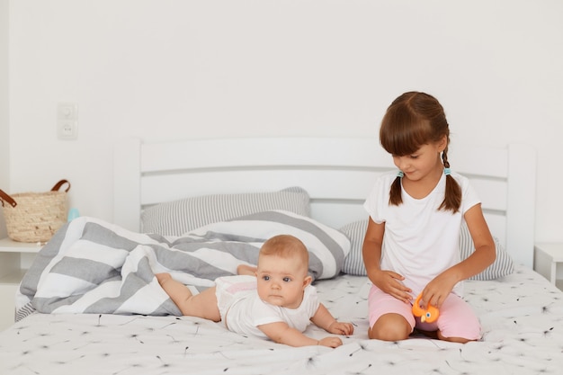 Cute dark haired child with pigtails sitting on bed near her infant sister, posing in light bedroom, elder girl looking at charming baby, spending time together at home.