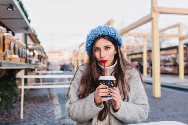 Cute dark-haired girl with sparkle manicure drinking tea on the street during winter photoshoot. Shy brunette young lady in trendy blue hat posing with cup of coffee in cold morning.