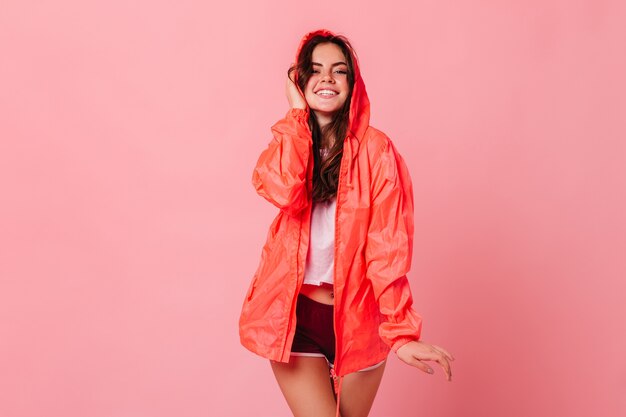 Cute dark-haired woman in white T-shirt and orange windbreaker laughs on pink wall