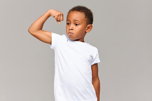 Cute dark skinned athletic male child with short Afro hairdo tensing arm muscle being proud of himself for working out. Confident cool sporty African boy demonstrating his strength at gray wall