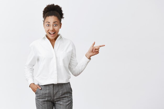 Cute dark-skinned female entrepreneur cannot believe she sees famous businesswoman during meeting, standing in trendy glasses and formal suit over gray wall, pointing right with forefinger