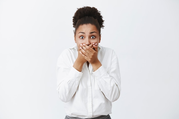 Free photo cute dark-skinned woman witnessed shocking and terrifying crime, covering mouth with palms not to shout, stooping, staring with popped eyes, frightened and stunned