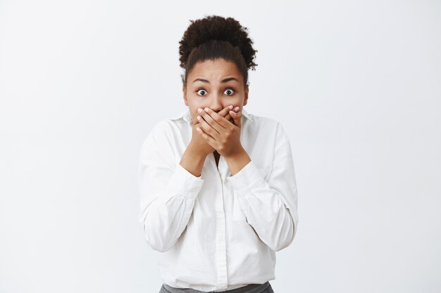 Cute dark-skinned woman witnessed shocking and terrifying crime, covering mouth with palms not to shout, stooping, staring with popped eyes, frightened and stunned