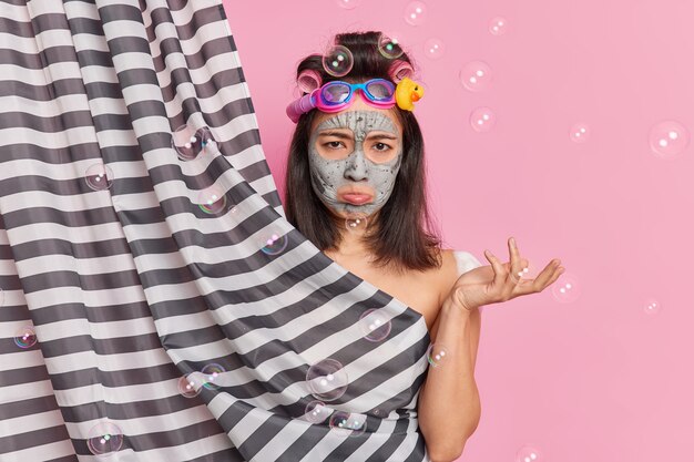 Cute displeased brunette woman undergoes beauty procedures at bathroom purses lower lip looks unhappily at camera applies clay mask and hair curlers poses behind shower curtain around bubbles