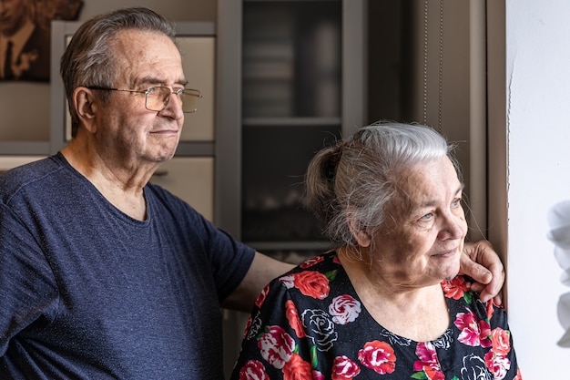 A cute elderly couple stands at the window and looks out for someone, waiting.