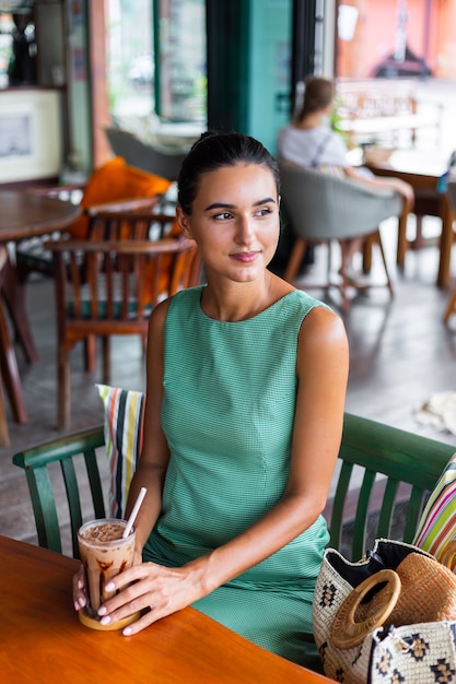 緑の夏のドレスでかわいいエレガントで落ち着いた幸せな女性は朝を楽しんでカフェでコーヒーと一緒に座っています
