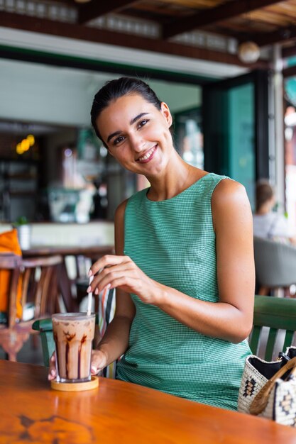 Cute elegant calm happy woman in green summer dress sits with coffee in cafe enjoying morning