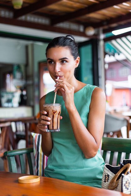 Cute elegant calm happy woman in green summer dress sits with coffee in cafe enjoying morning