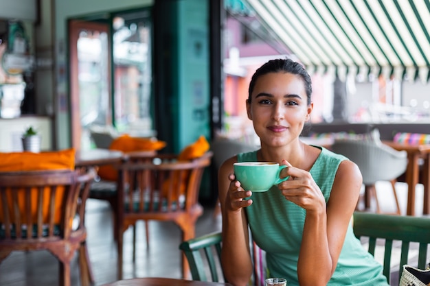 Cute elegant calm happy woman in green summer dress sits with coffee in cafe enjoying morning