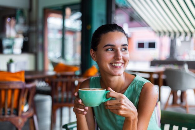 Cute elegant calm happy woman in green summer dress sits with coffee in cafe enjoying morning