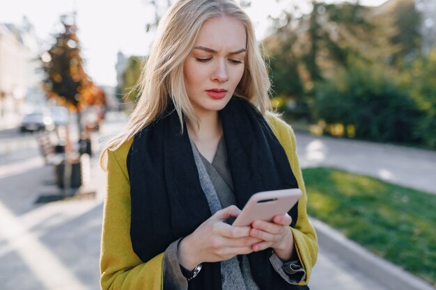 Cute emotional attractive blonde woman in coat with smartphone walks the city street