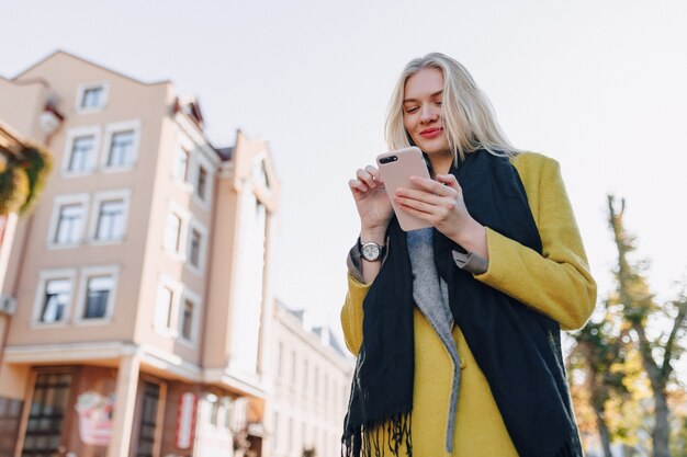 スマートフォンでコートを着たかわいい感情的な魅力的なブロンドの女性が街を歩く