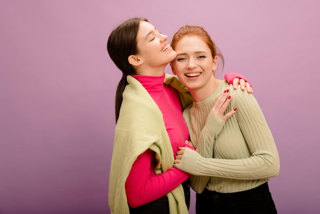 Cute european girls brunette and redhead in casual clothes stand hugging smiling on purple background People emotions lifestyle and fashion concept