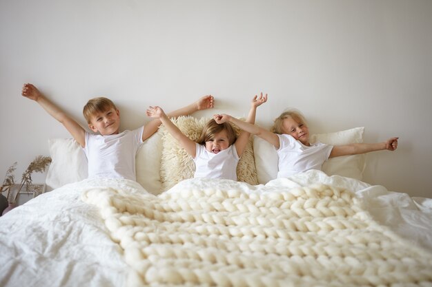 느린 게으른 아침을 즐기고 부모의 침실에서 스트레칭을하는 귀여운 유럽 형제. 세 명의 사랑스러운 캐주얼 옷을 입은 아이들이 침실에서 함께 누워서 팔을 뻗고 일어나기를 꺼림
