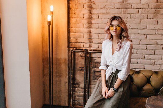 Cute fair-haired girl in white shirt sitting on leather sofa in room with loft design and looks with interest