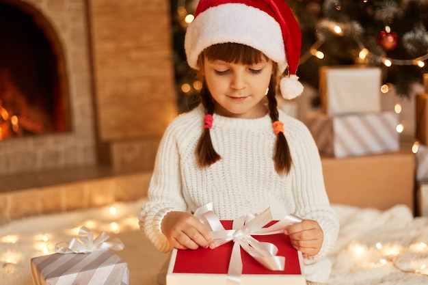 산타클로스의 선물 상자를 여는 귀여운 여자 아이는 흰색 스웨터와 산타클로스 모자를 쓰고 벽난로와 크리스마스 트리가 있는 축제 공간에서 포즈를 취합니다.