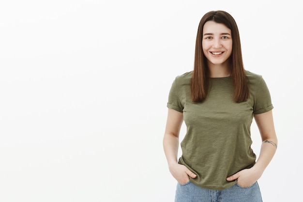 Cute female coworker with ambitious gaze holding hands in pockets waiting for assignment, smiling broadly feeling relaxed and happy over gray wall wearing olive t-shirt