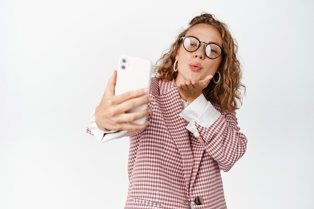 Симпатичная женщина-менеджер, делающая селфи, отправляющая поцелуи в камеру мобильного телефона, романтический видеозвонок в приложении smarpthone, стоящая на белом