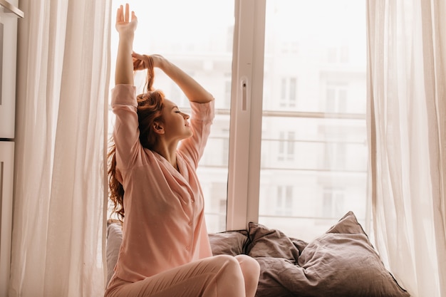 Cute female model in pink pajama enjoying morning. Pleasant ginger woman sitting on bed.