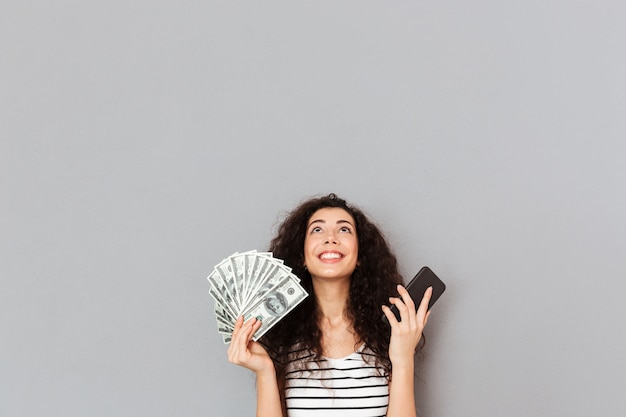 Cute female in striped t shirt holding fan of 100 dollar bills and cell phone in hands looking up being thankful cannot believe in her triumph