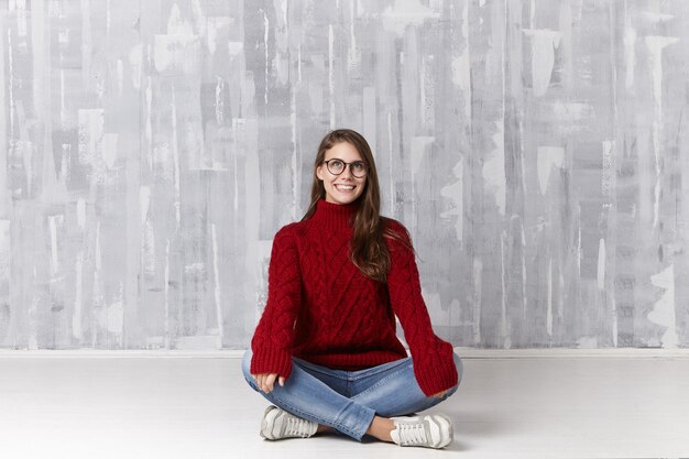 Cute female teenager wearing stylish glasses, sneakers, jeans and warm pullover sitting on floor with legs folded, looking up and smiling joyfully