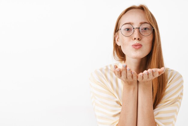 cute feminine redhead female in trendy glasses with natural freckles folding lips and blow kiss expressing love and passion