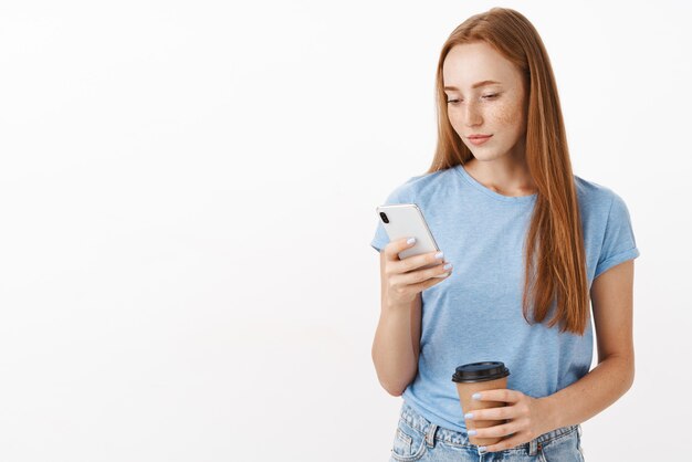 파란색 티셔츠에 귀여운 여성 빨간 머리 여자는 장치 화면을보고 커피와 전화 메시징 종이 컵을 들고 스마트 폰에 메모를 작성하는 데 집중되고