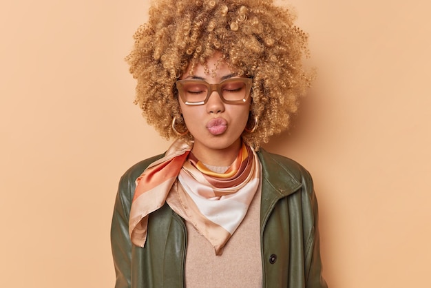 Cute flirty young woman folds lips waits for kiss sends lovely mwah feels romantic wears leather jacket spectacles keeps eyes closed isolated over beige background expresses love and tenderness